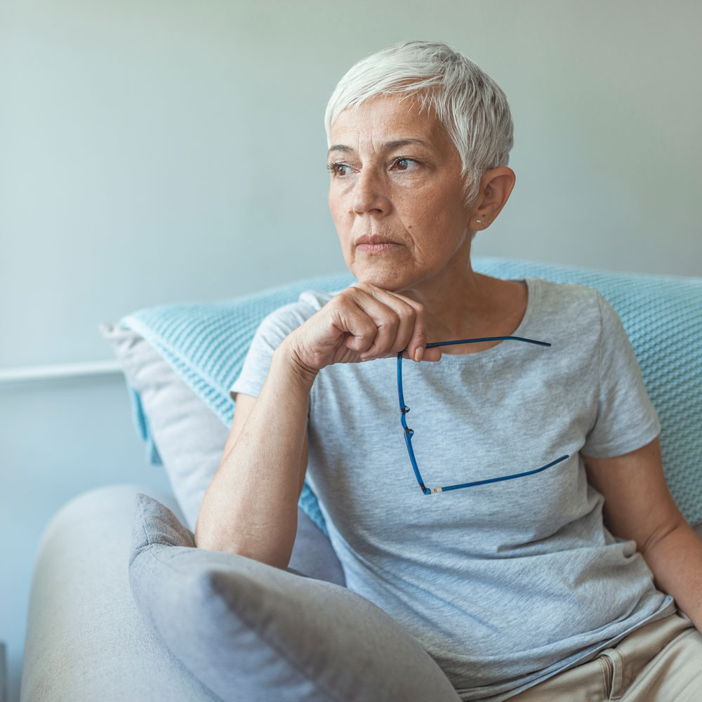 Older woman with short gray hair sits on a couch, holding her glasses, and looks thoughtfully into the distance.