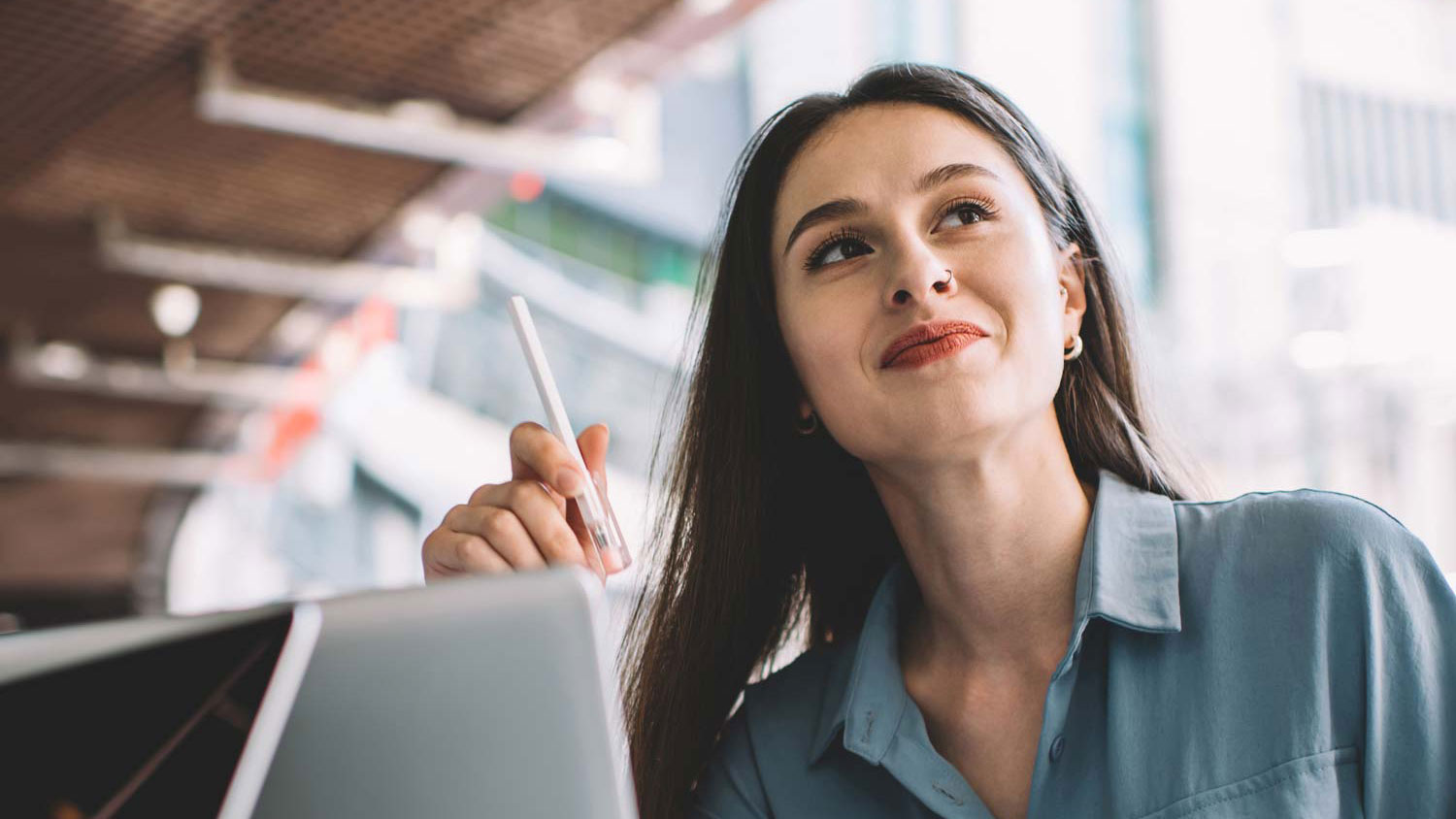 Young woman holding a pen and smiling thoughtfully while sitting in front of a laptop.