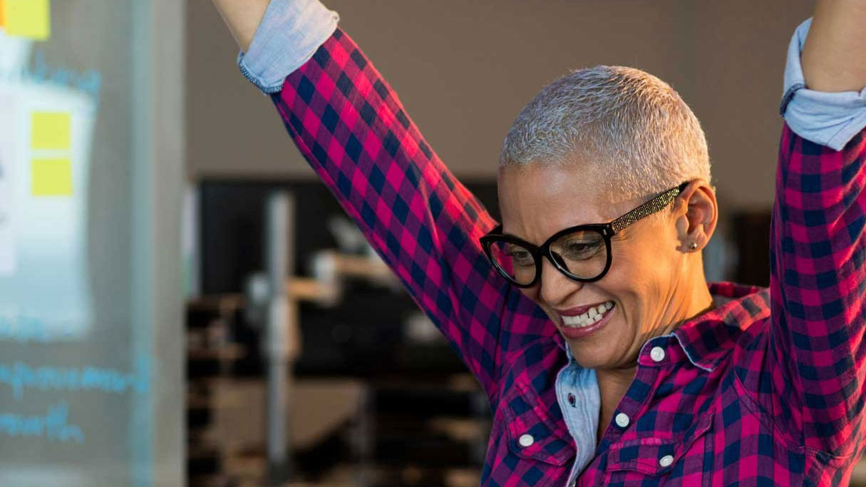 Smiling person with short gray hair and glasses raises arms in celebration, wearing a pink plaid shirt.