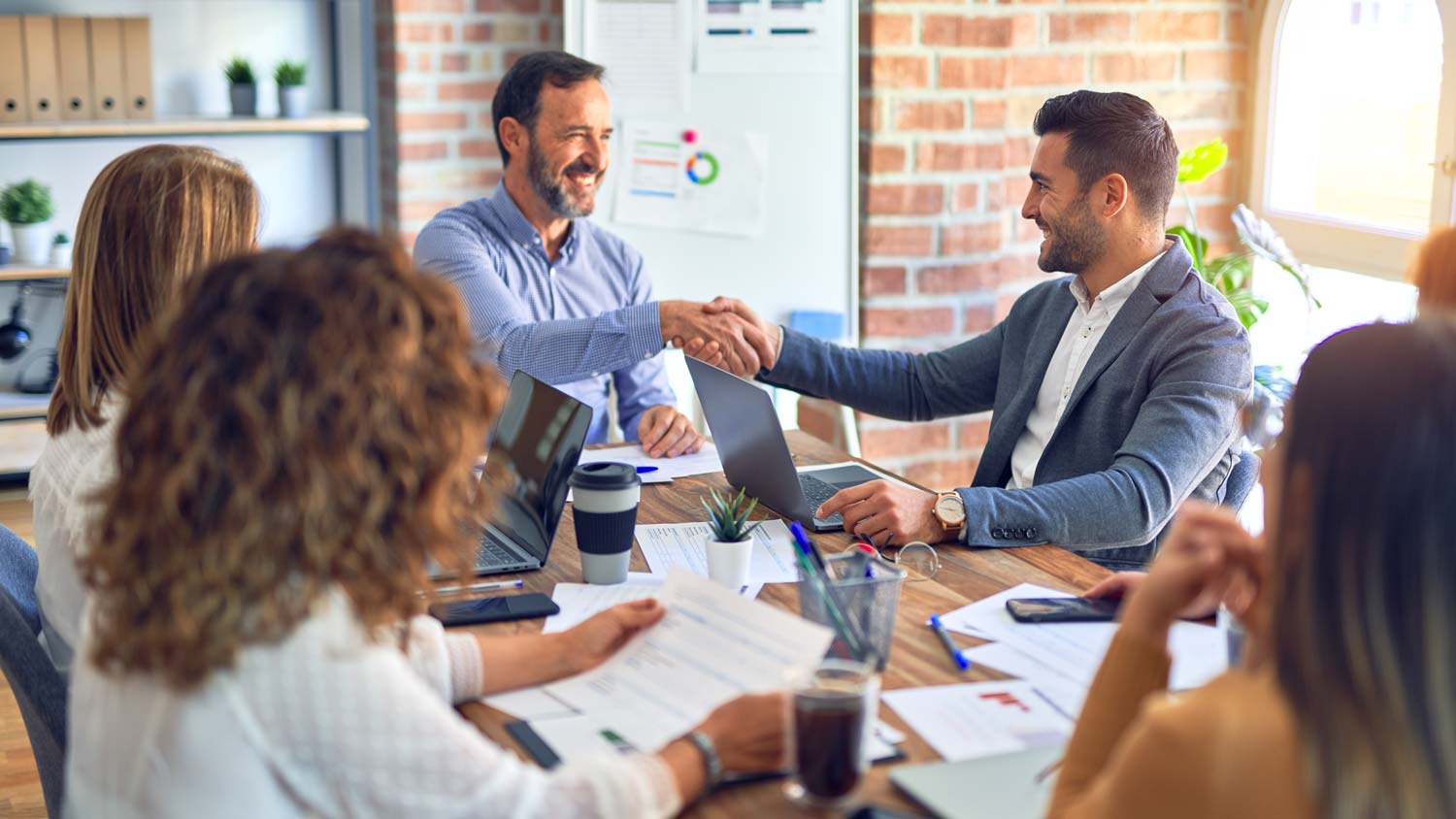 Two businessmen shake hands at a meeting table with colleagues, laptops, and documents in a bright office.