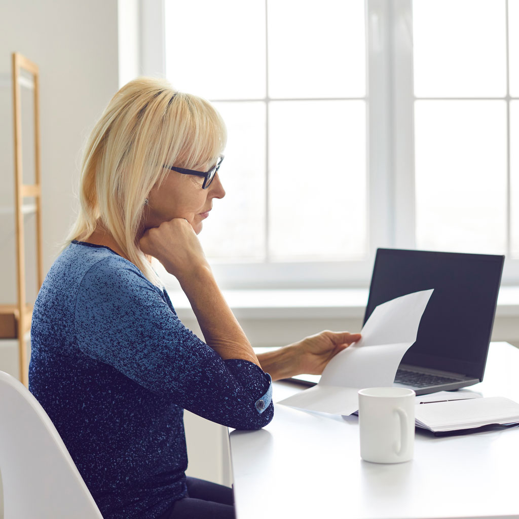 Woman with blonde hair and glasses reading papers at a desk with a laptop and coffee mug.